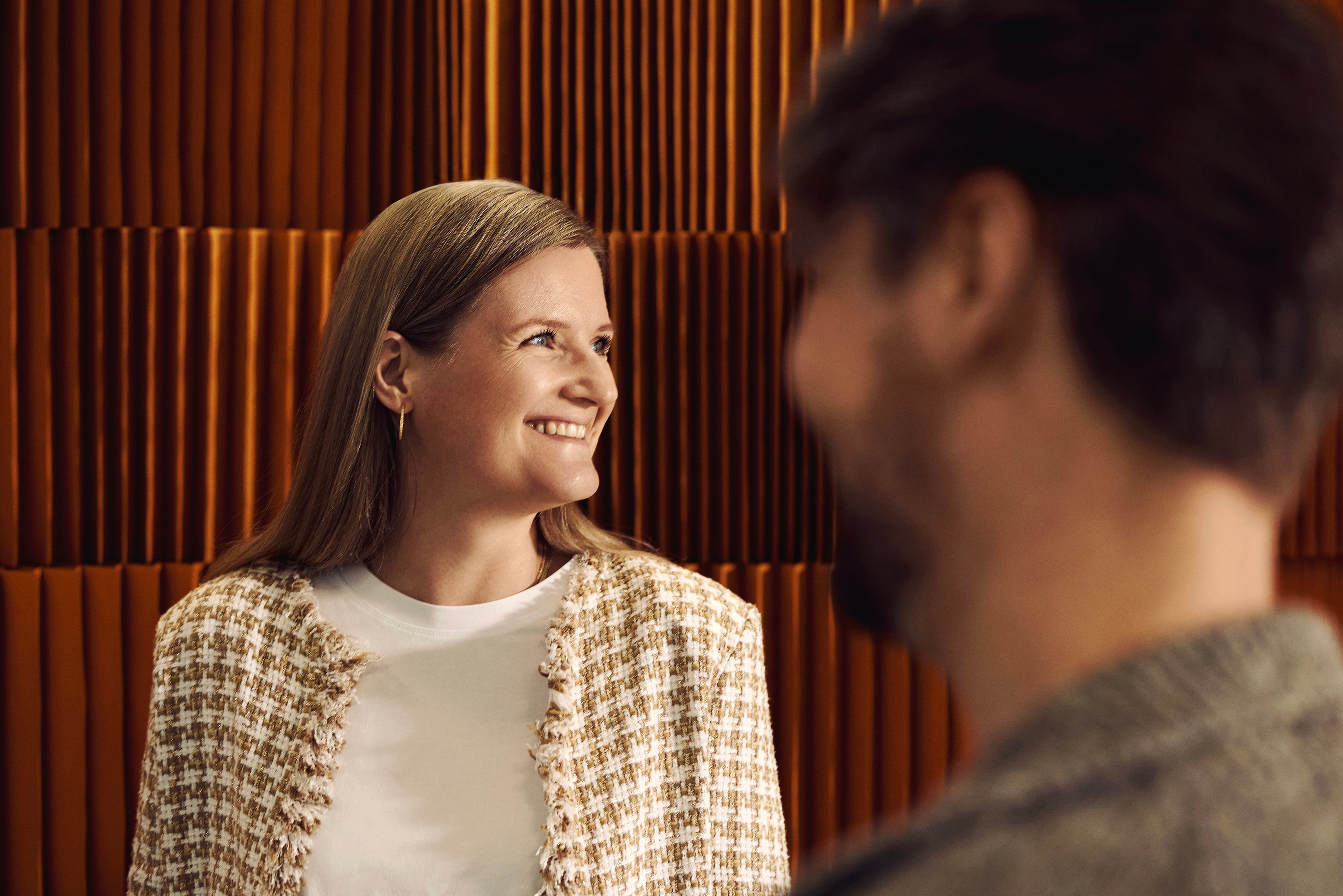 To personer i samtale foran orange vægpanel. Den ene smiler og ser mod siden