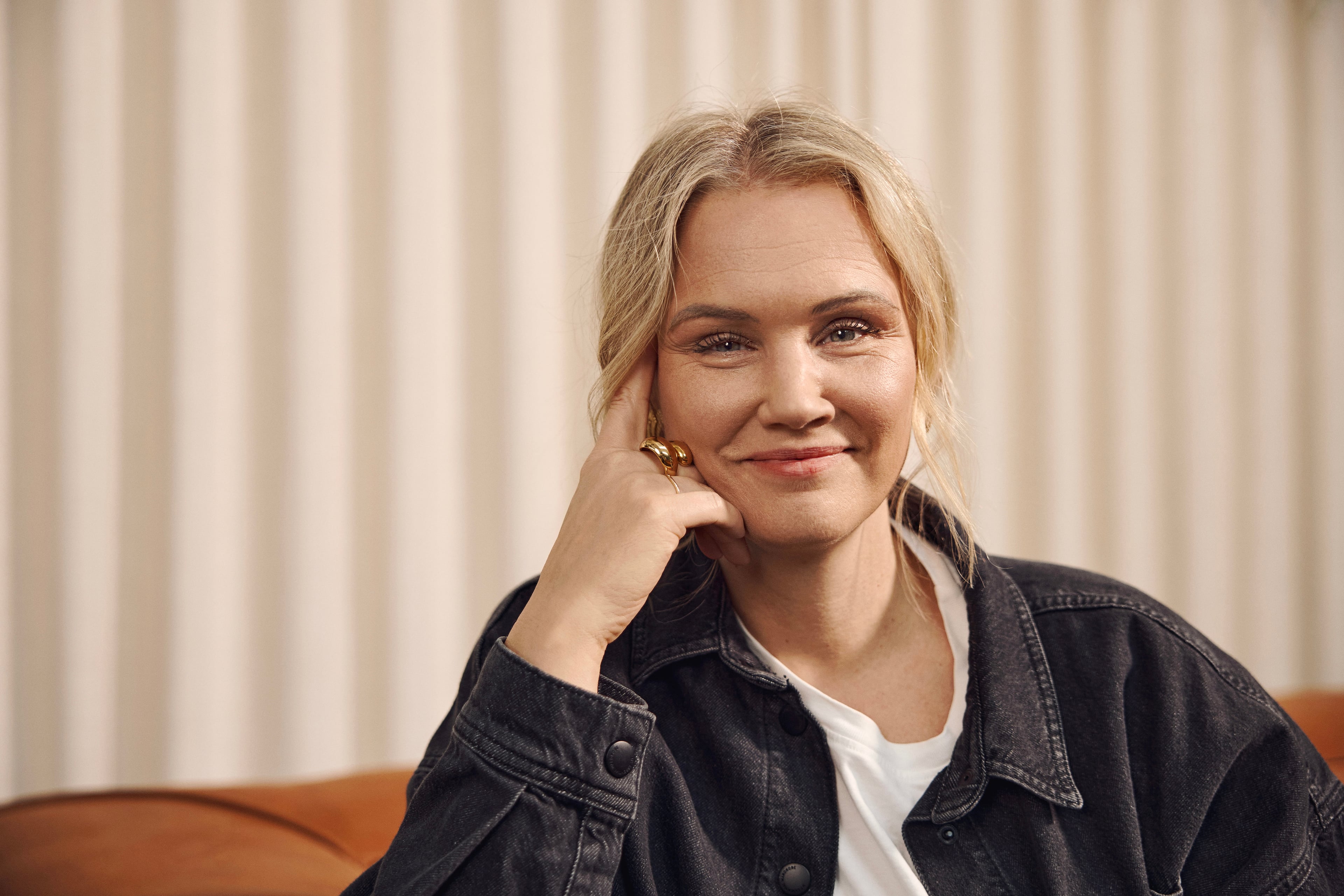 Kvinde med blondt hår i denimjakke sidder foran lysere gardin og smiler