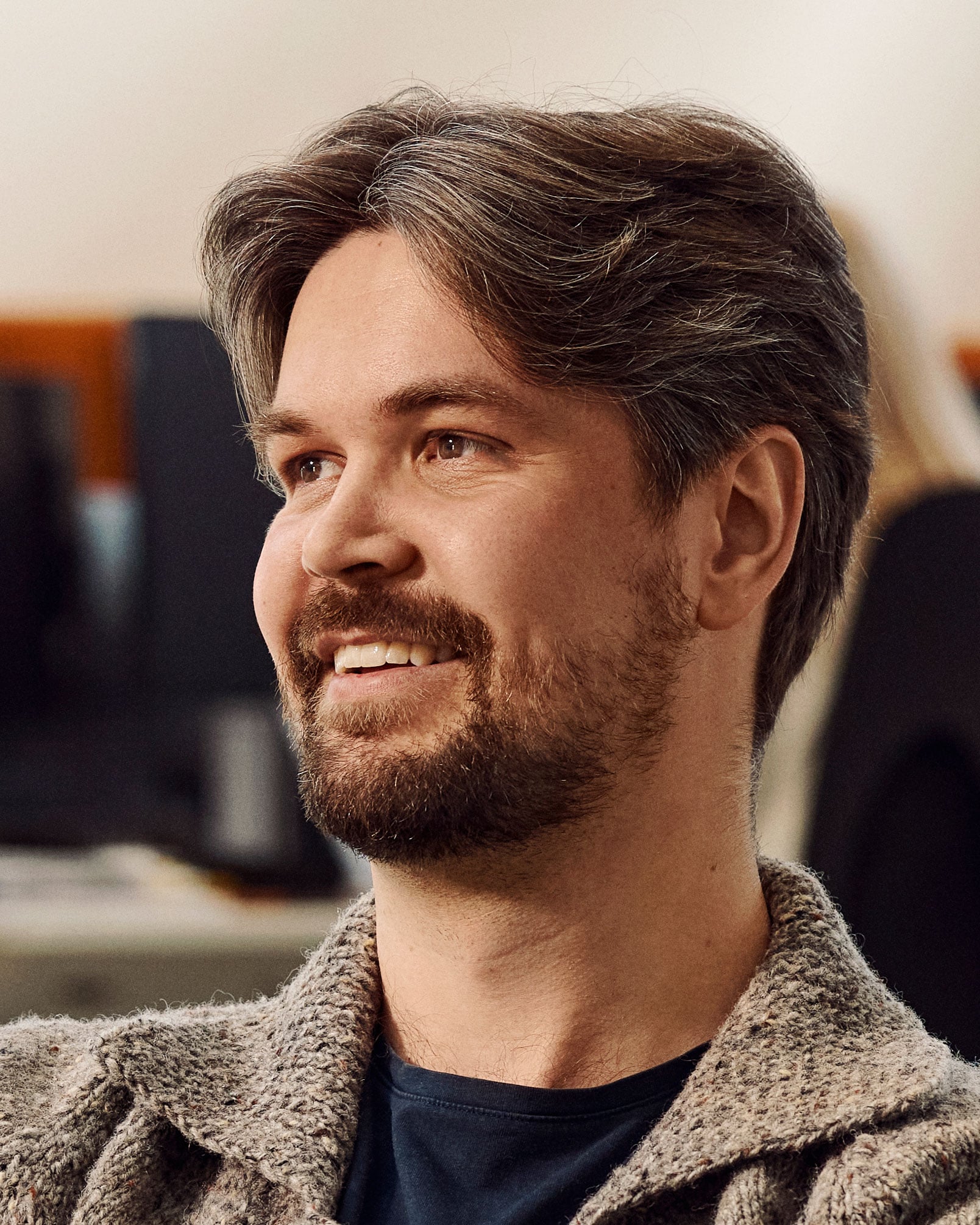 Mand med mørkt hår og skæg, iført grå cardigan, smiler mod siden